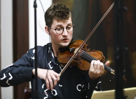 A photograph. A string concert in Villa Decius. A man in a blue shirt plays the violin.